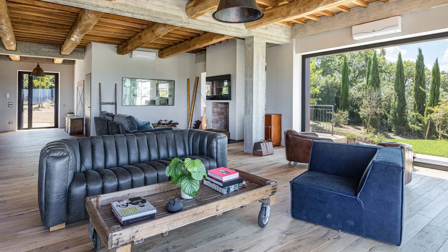 A spacious living room has leather sofas, a rustic coffee table, exposed wooden beams, and a large garden-facing window.