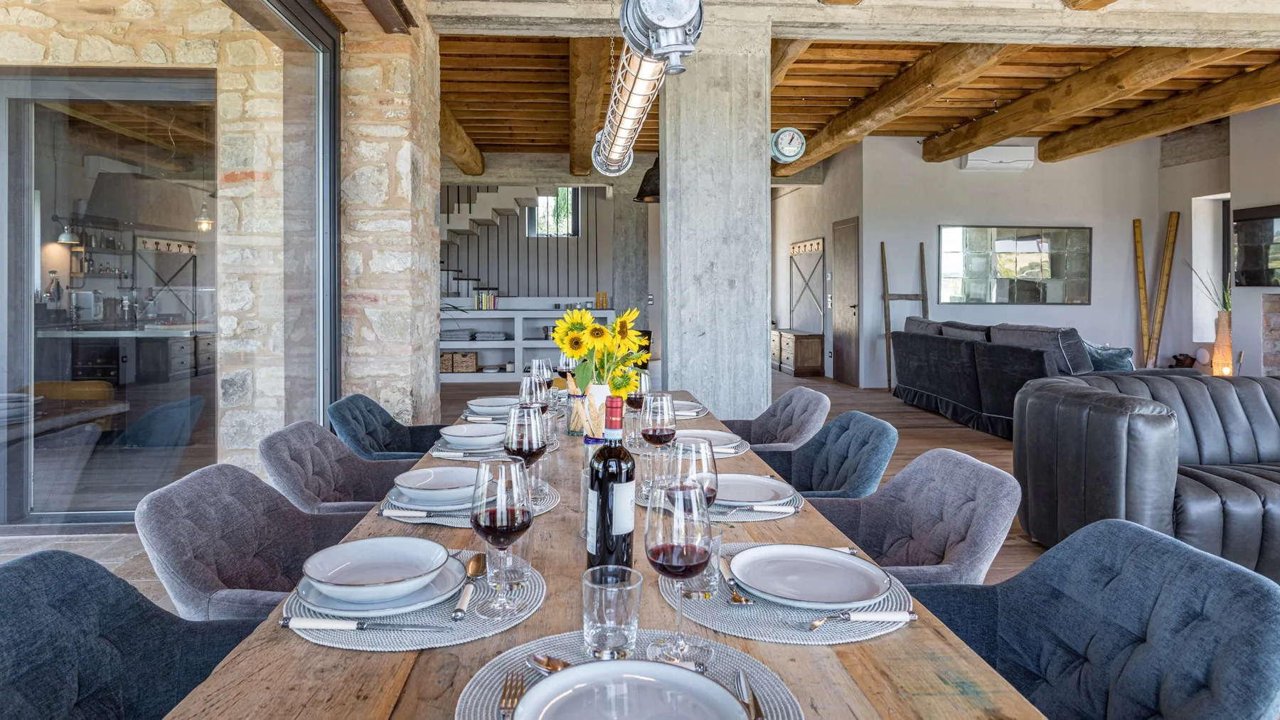 A long dining table is set with plates and wine glasses beside a living area with gray sofas.
