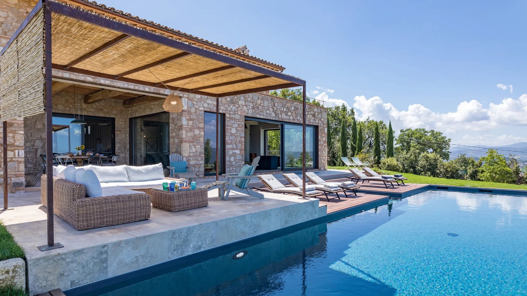 A stone house opens onto a patio with cushioned seating, loungers, and a clear blue pool.