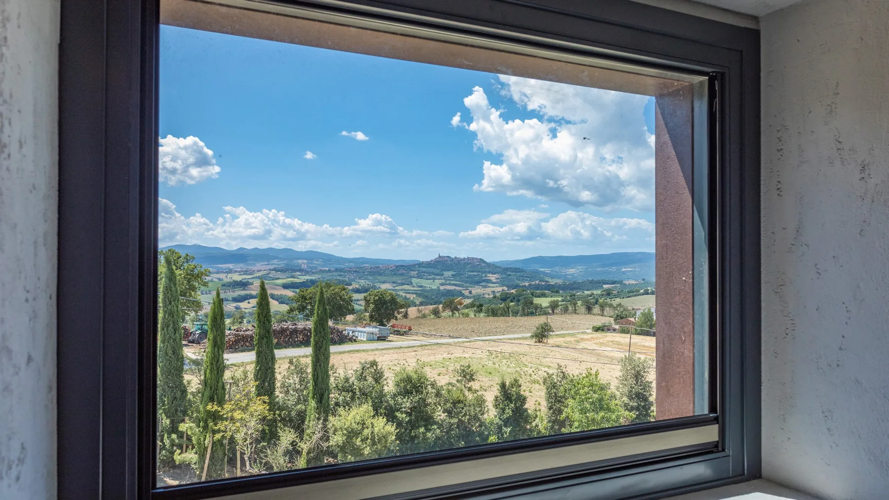 A large window frames a rural landscape with cypress trees, fields, distant hills, and a bright blue sky.