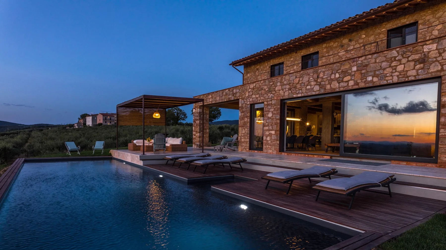 A stone house overlooks a swimming pool and wooden deck with loungers at dusk.
