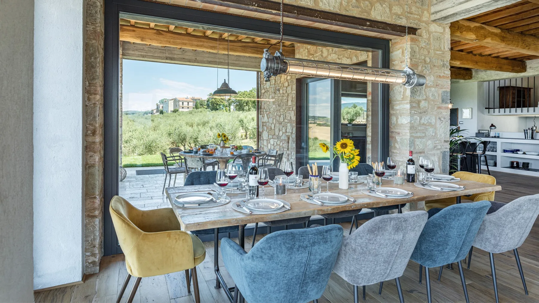 A stone-walled dining area features a long set table, upholstered chairs, and large windows opening onto greenery.
