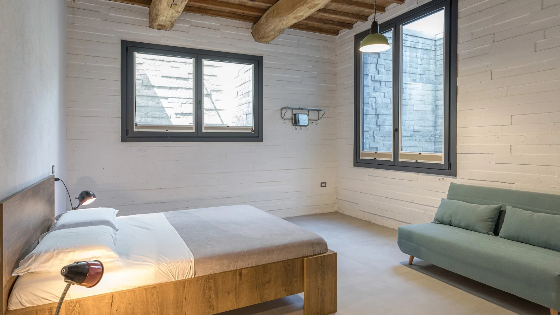 A bright bedroom with a wooden bed, pale bedding, a green sofa, and exposed ceiling beams.