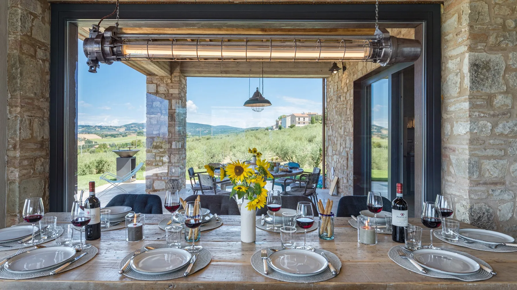 A stone dining terrace is set with plates, wine glasses, and sunflowers, overlooking a green countryside landscape.