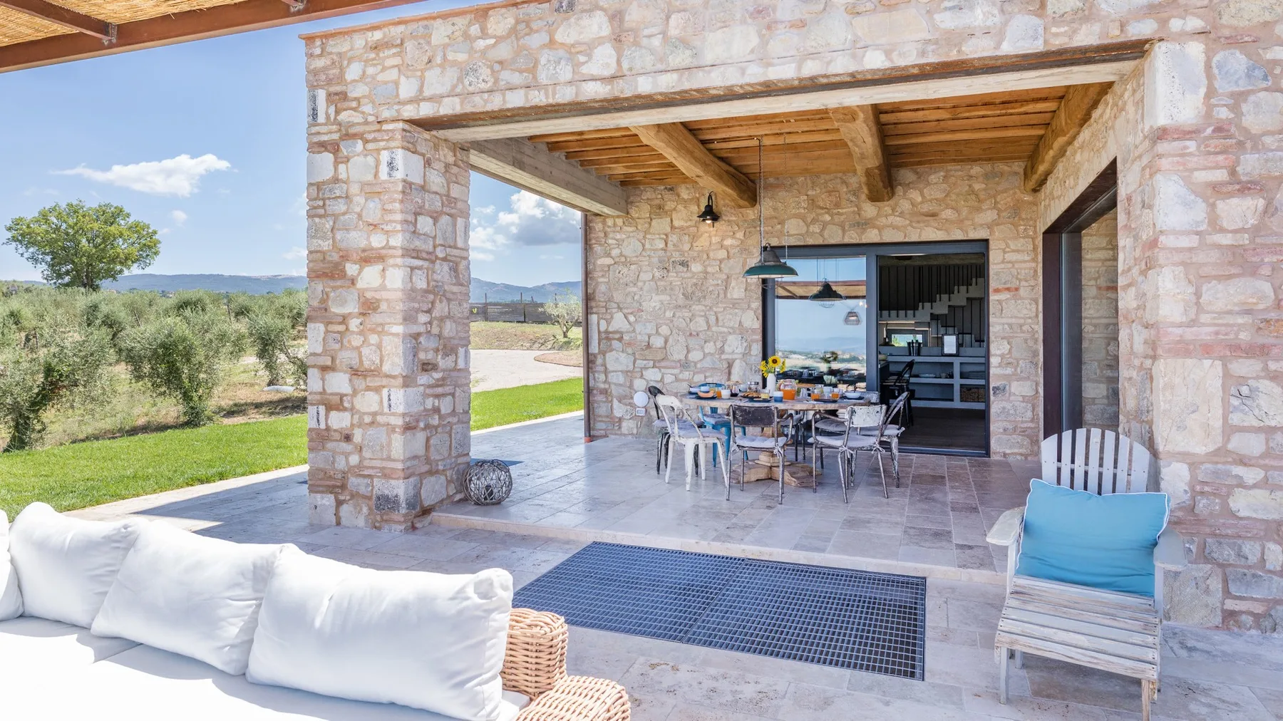 A stone terrace with a wooden ceiling, outdoor dining table, lounge seating, and views of the countryside.
