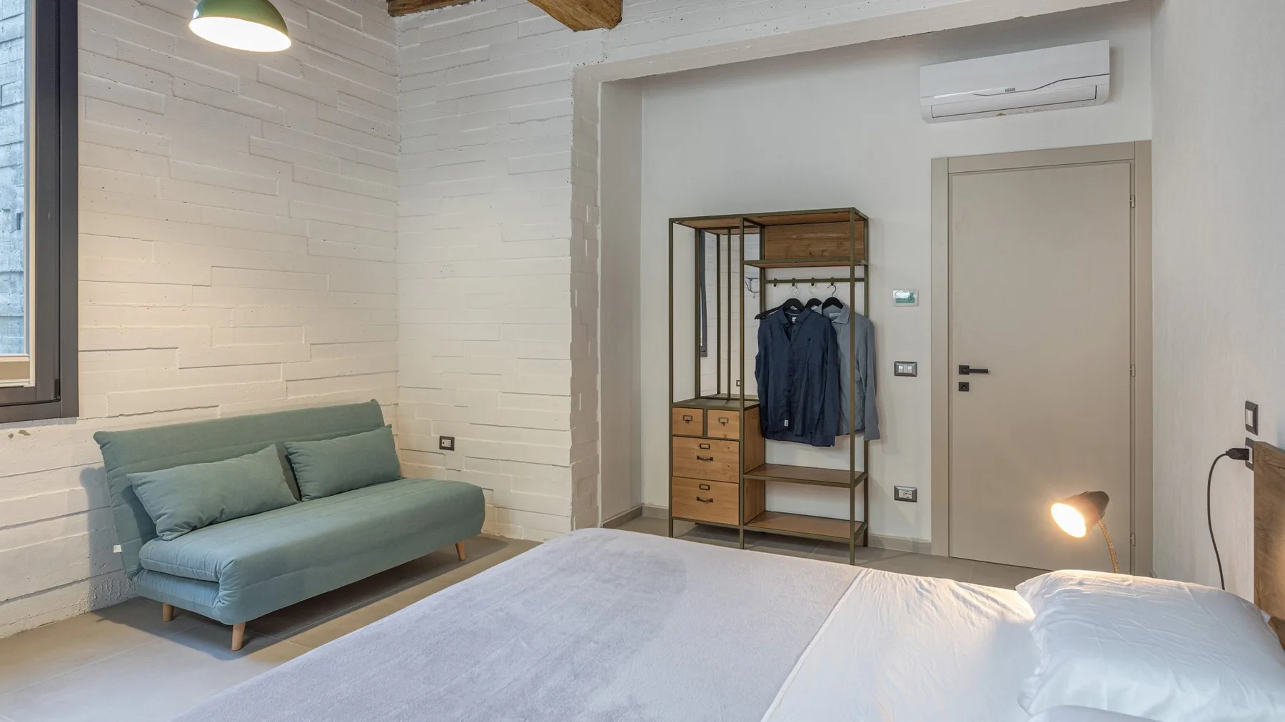A bedroom with a neatly made bed, a pale blue sofa, and an open wardrobe beside the door.
