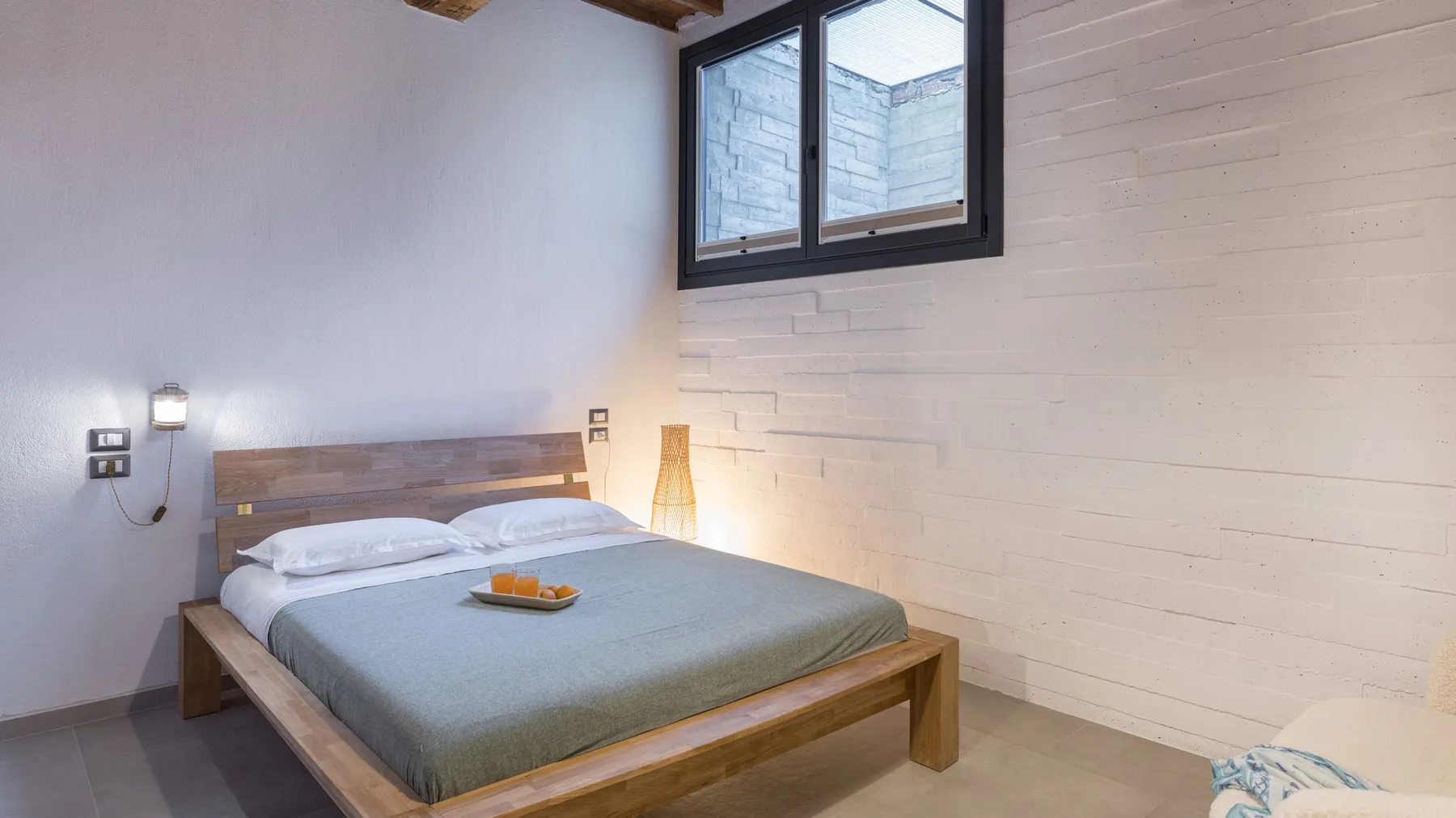 A simple bedroom with a wooden bed, gray bedding, and a small high-set window.