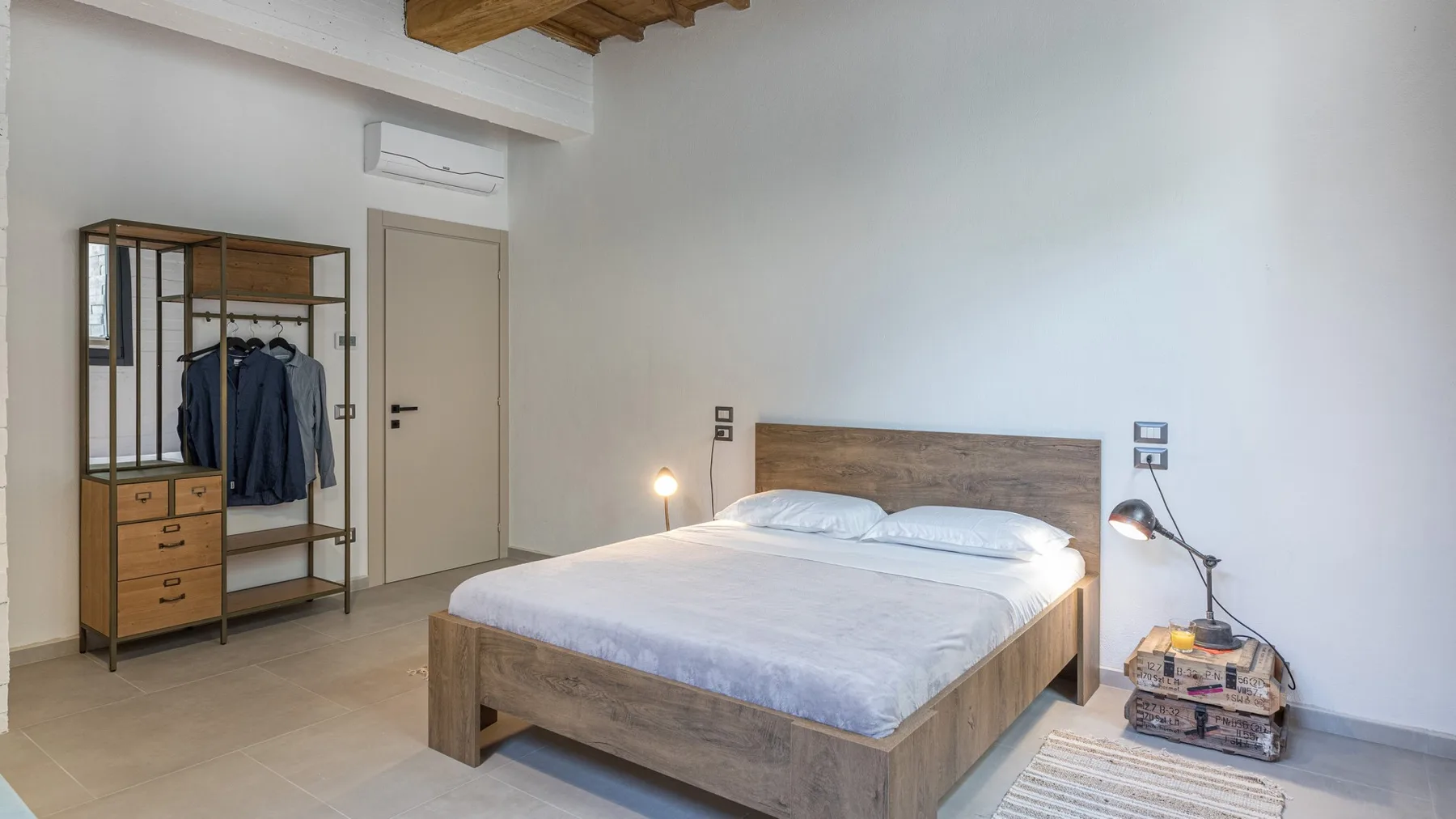 A bright bedroom has a wooden bed with white bedding, a wardrobe rack, and bedside lamps.