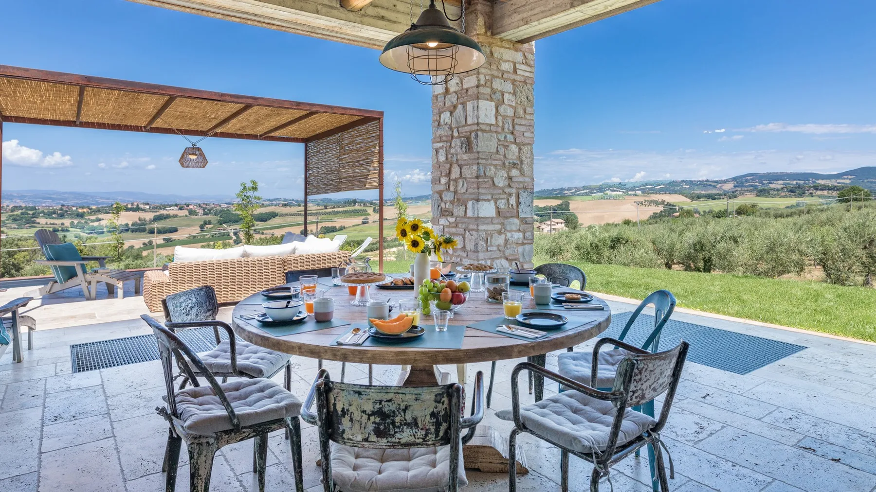 A covered stone patio features a round dining table, wicker seating, and views across open countryside.