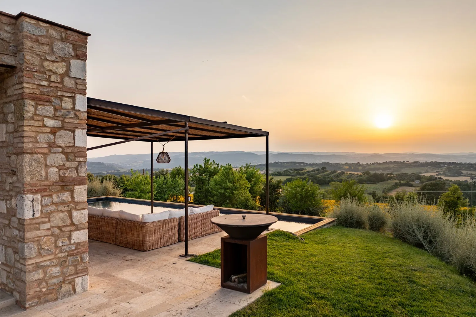 A stone patio with a pergola, wicker seating, and a fire pit overlooks rolling hills at sunset.