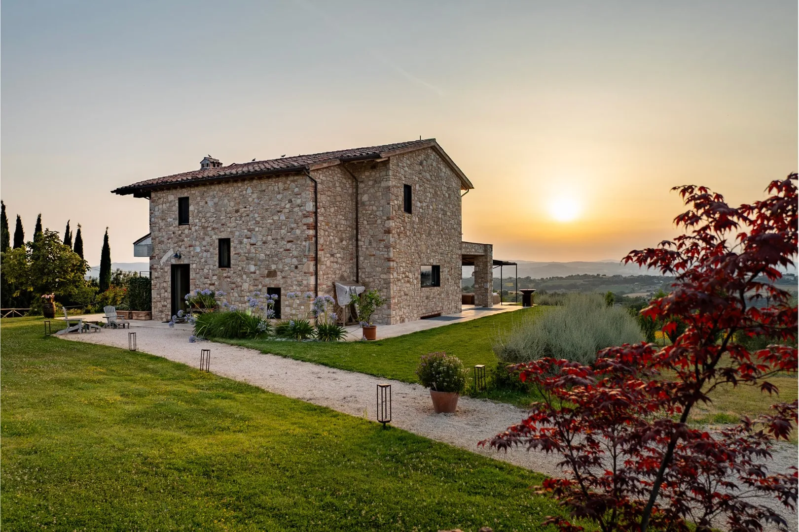 Stone house surrounded by a lawn and garden, with a gravel path at sunset.