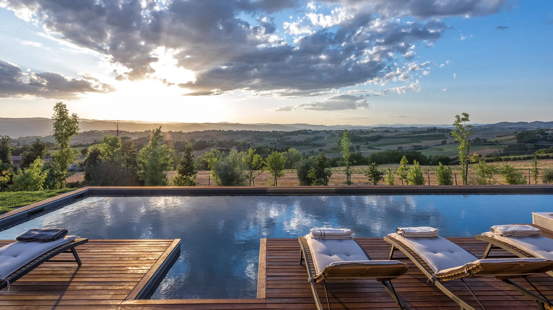 Outdoor pool with wooden sun loungers overlooks a green landscape beneath a cloudy sunset sky.