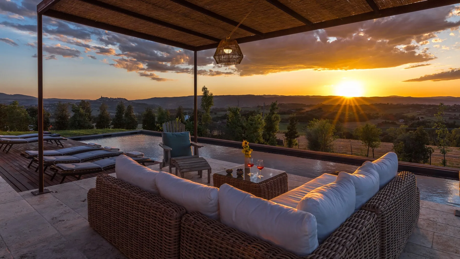 A covered outdoor lounge has wicker seating, cushioned chairs, and a sunset view over the hills.