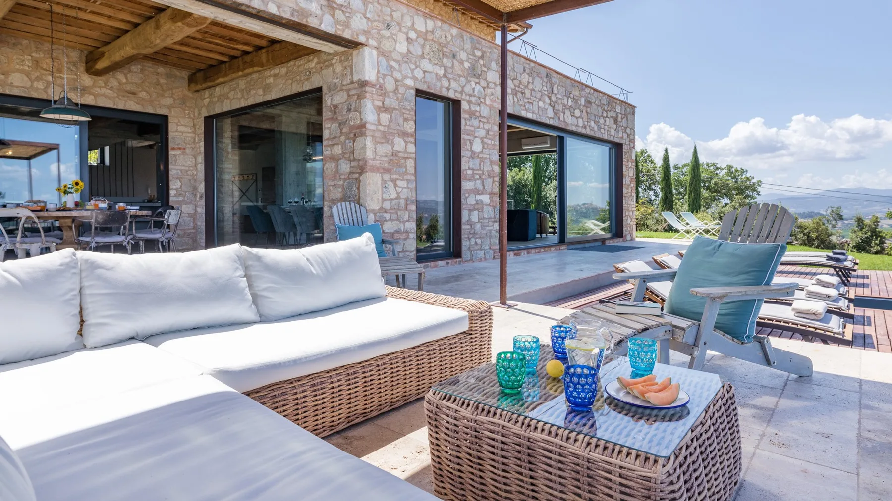 A stone house opens onto a sunny terrace with white wicker seating, a coffee table, and a pool.