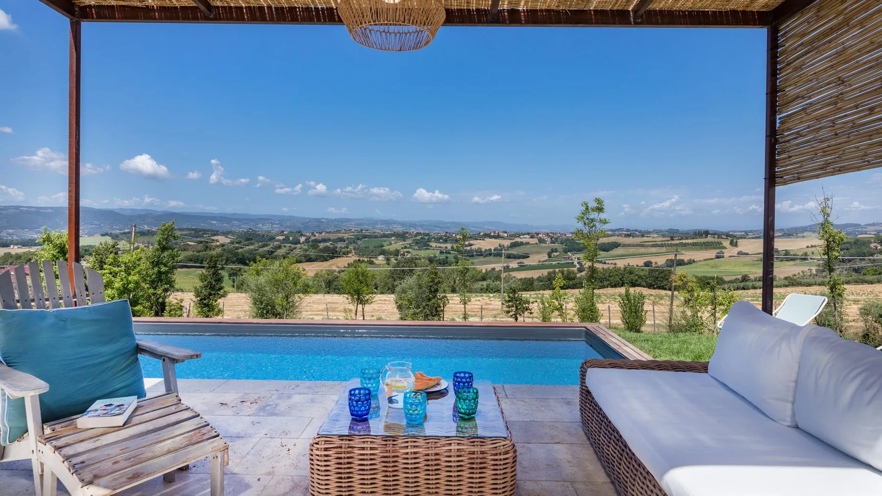 A shaded patio overlooks a swimming pool and rolling countryside, with cushioned loungers and a low table.