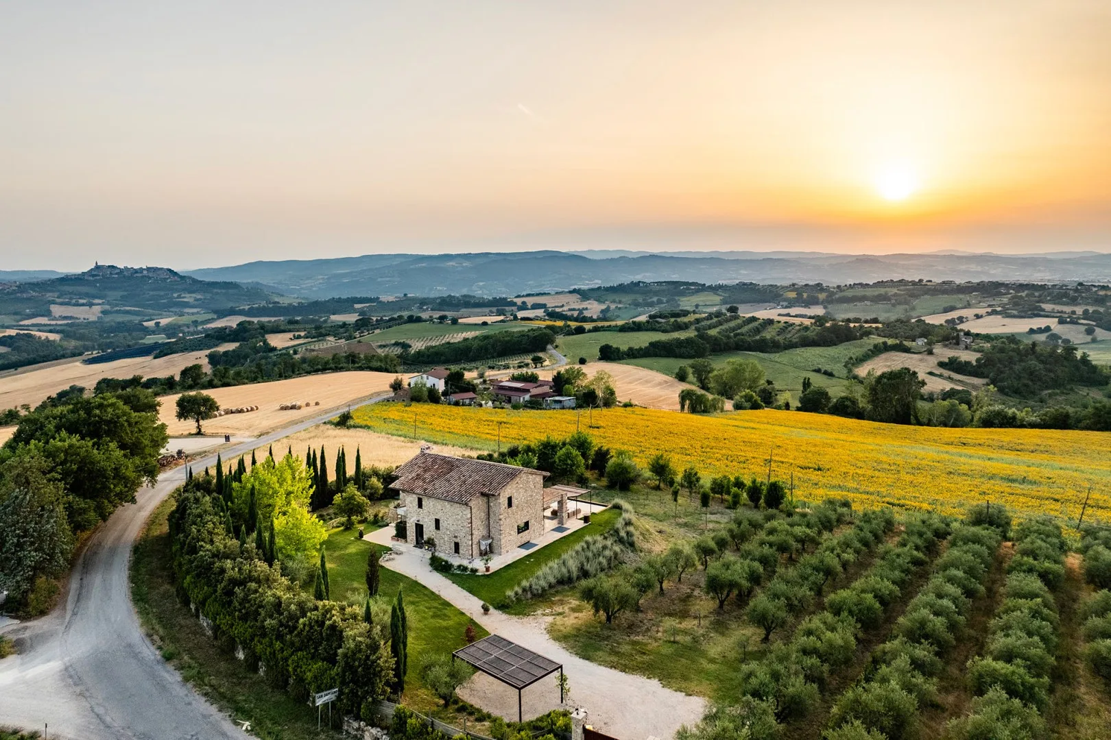 A stone farmhouse sits among orchards and bright fields, with rolling hills beneath a low setting sun.