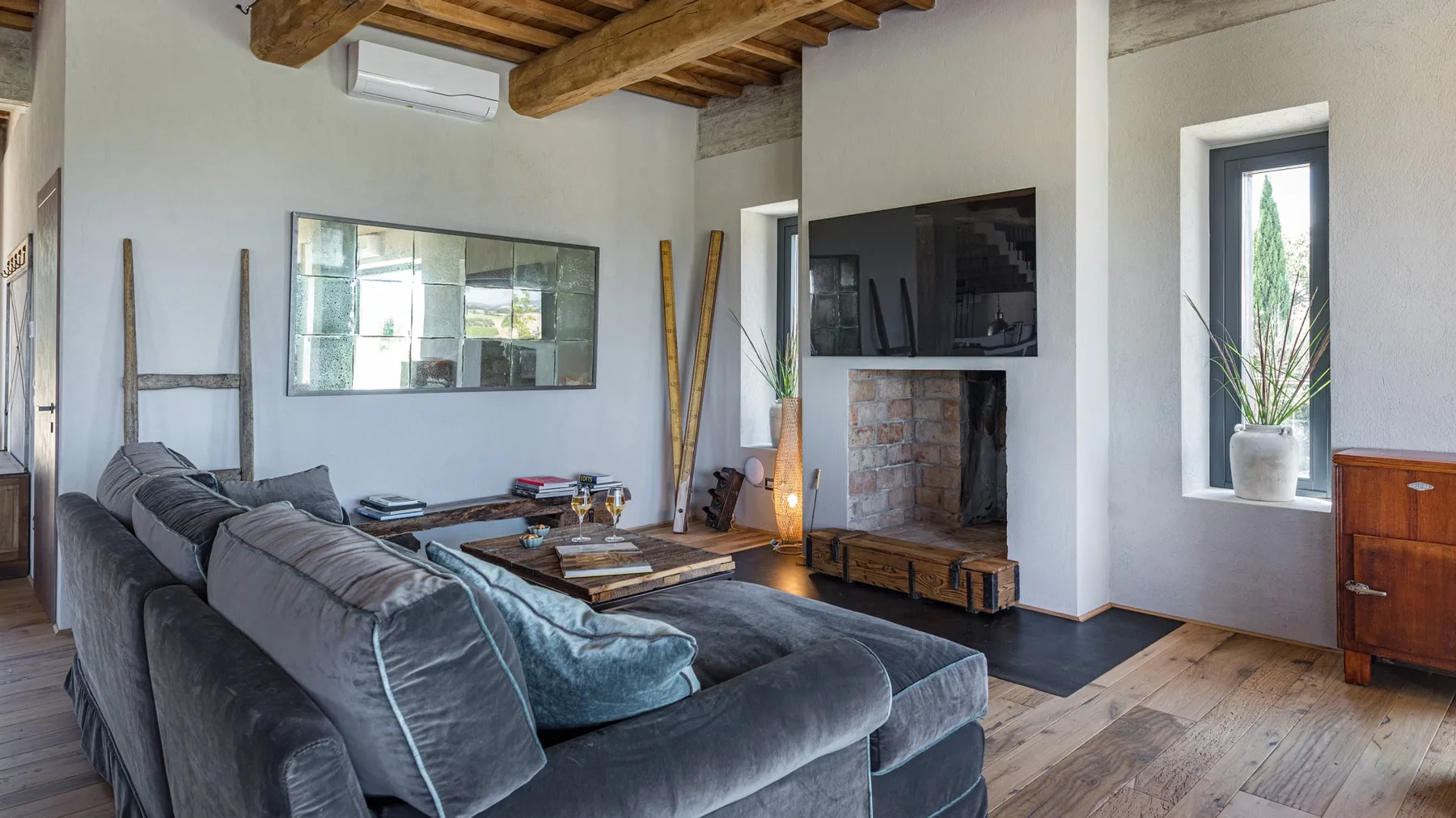 A living room with a large gray sectional sofa, a brick fireplace, and exposed wooden ceiling beams.