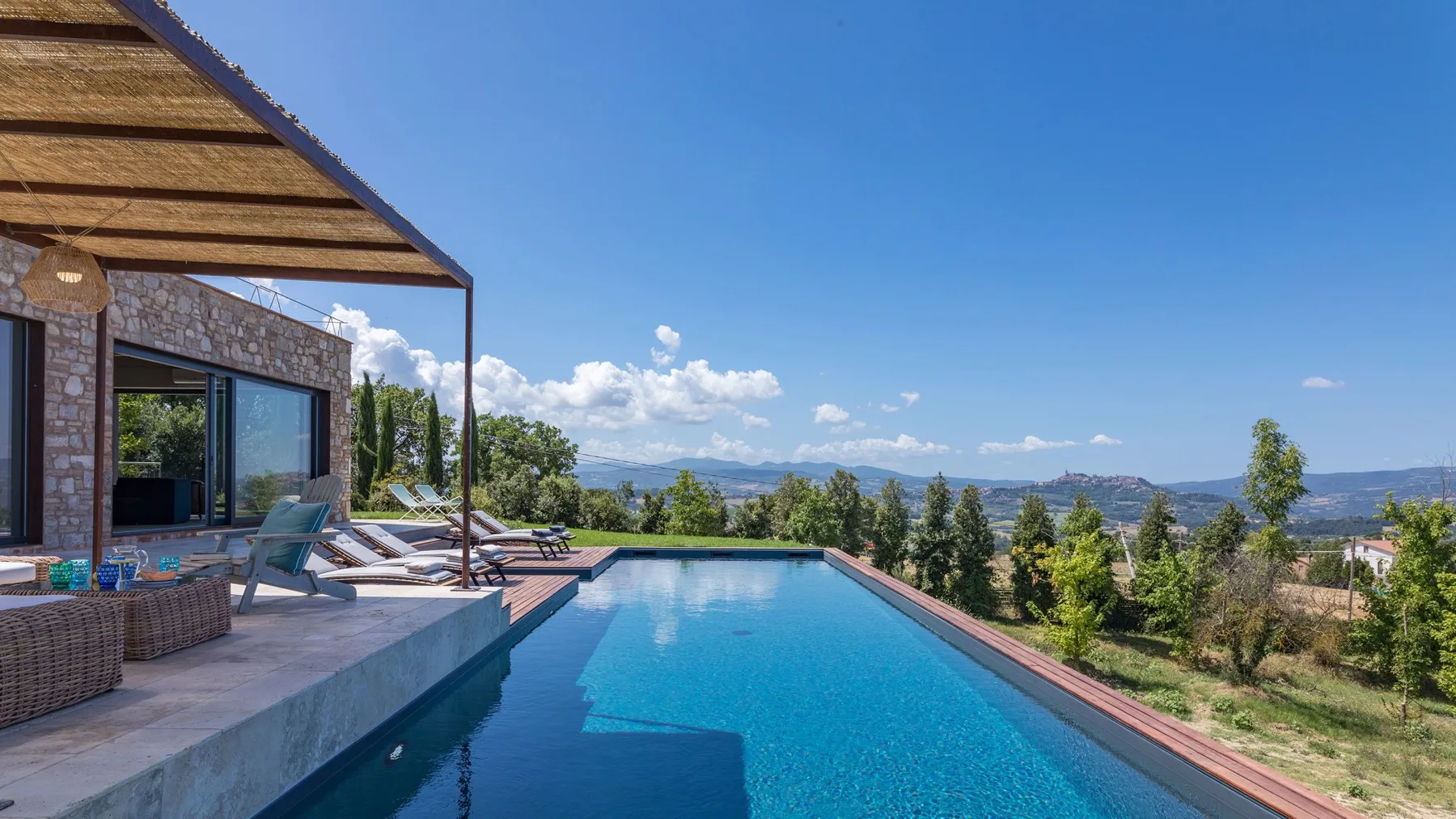 A long blue swimming pool borders a stone patio with loungers beneath a wooden pergola.