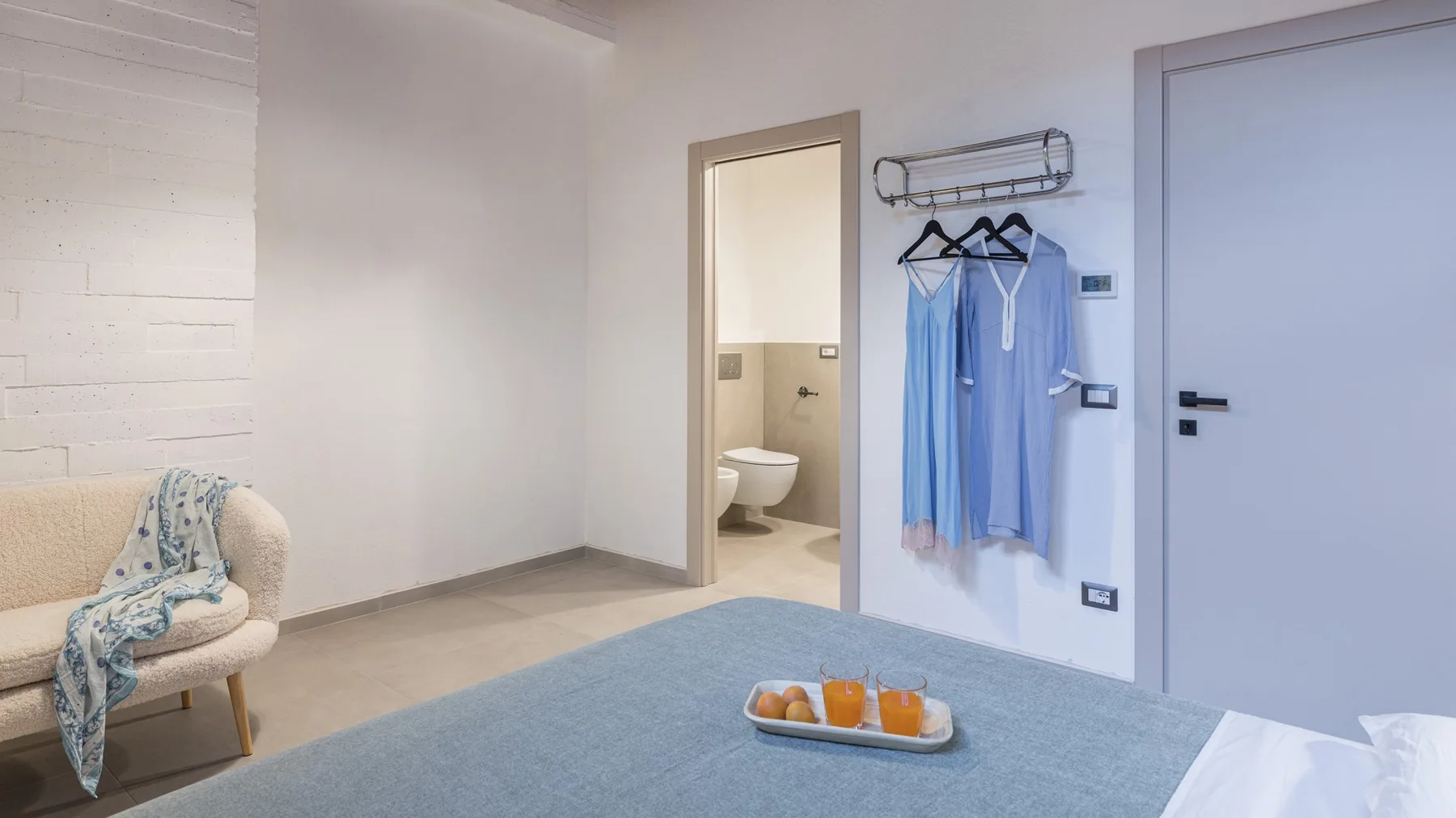 A bright bedroom has a blue-covered bed, a cream armchair, hanging blue robes, and an open bathroom doorway.