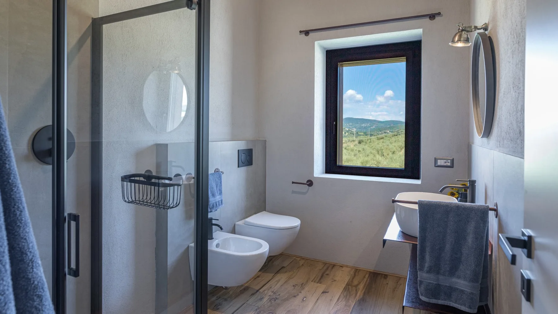A modern bathroom with a glass shower, wall-mounted toilet, sink, and window overlooking green hills.