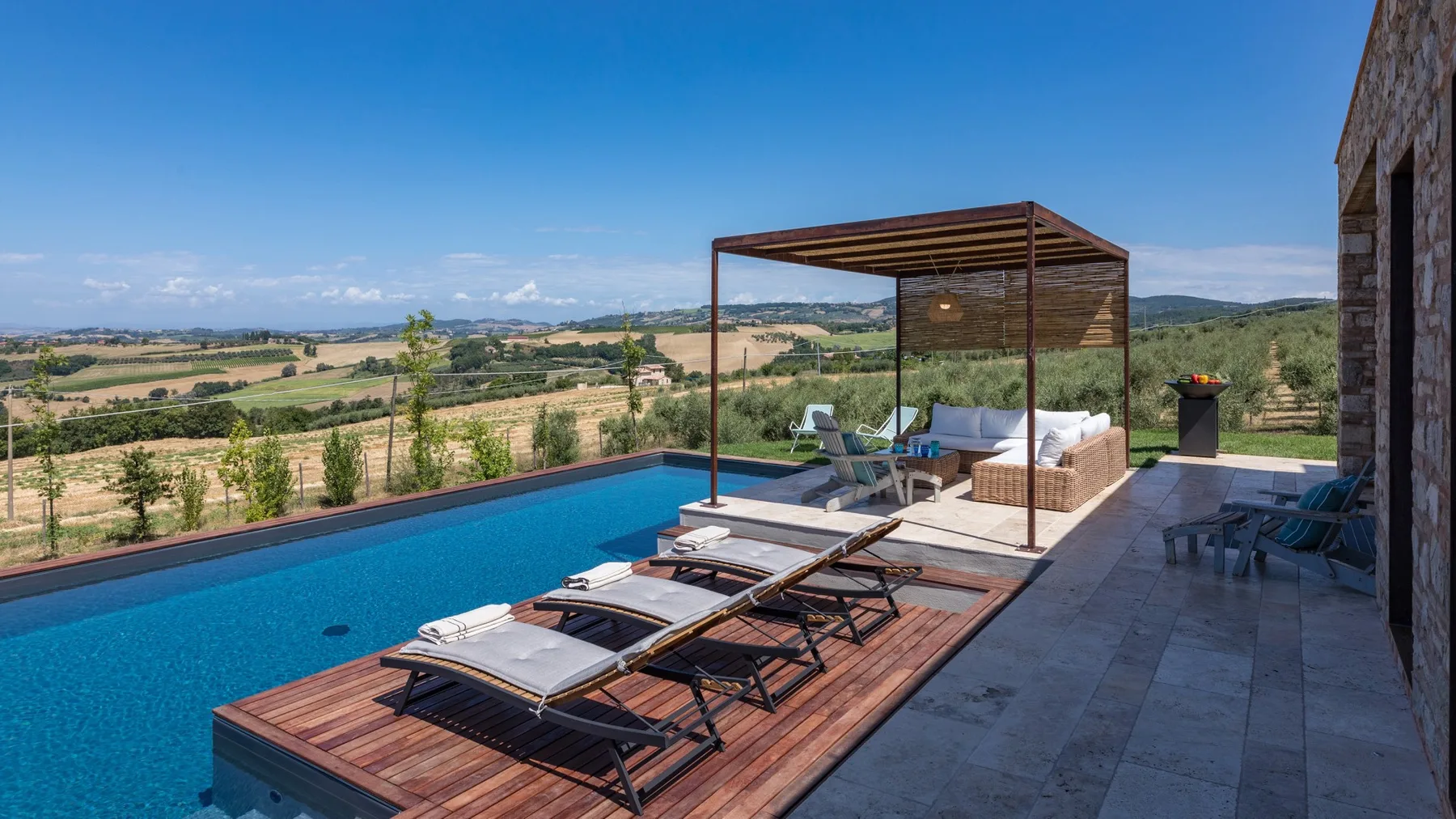 An outdoor pool overlooks rolling countryside, with wooden sun loungers and a shaded seating area.
