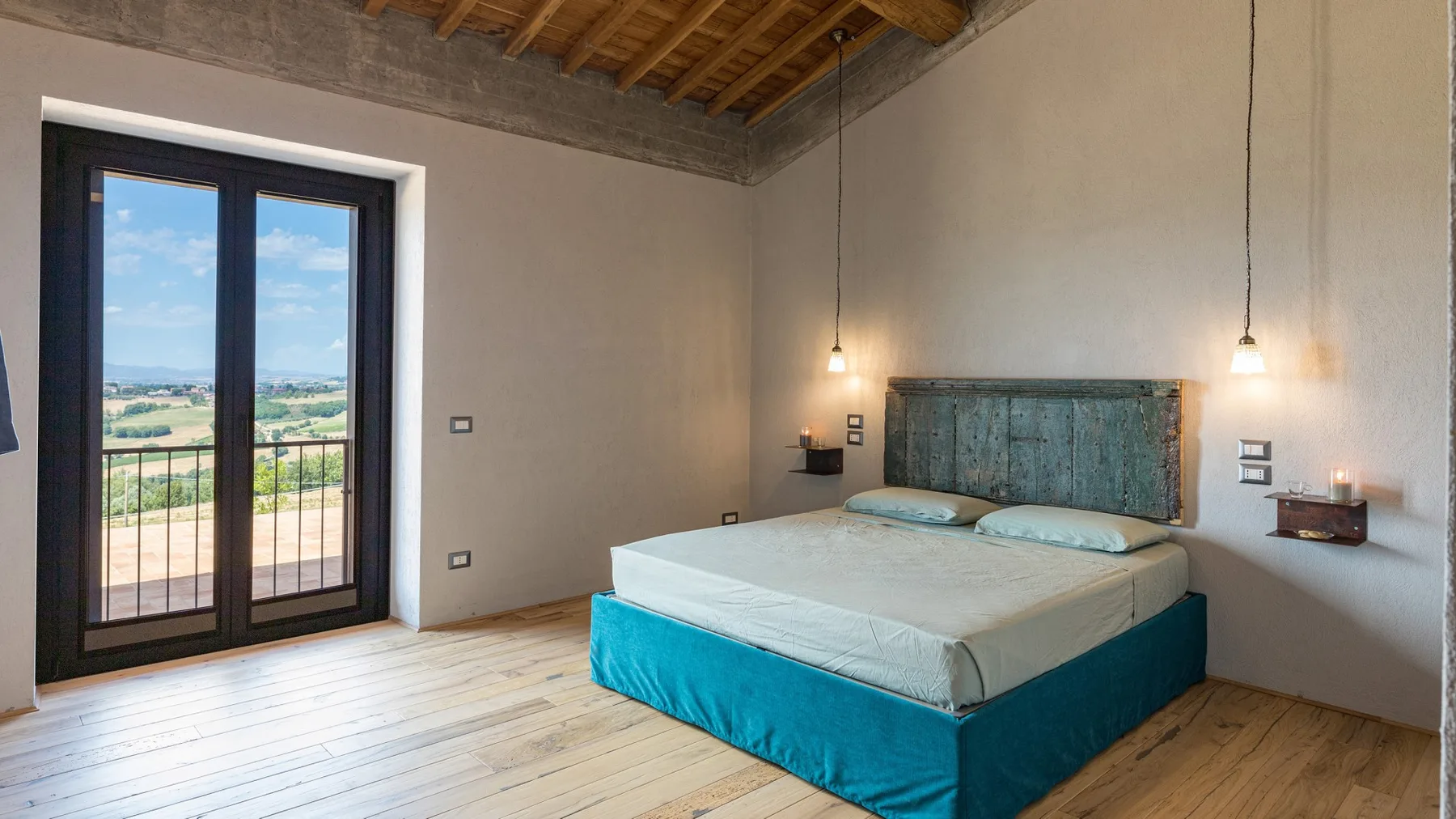 A bedroom with a double bed, teal bed skirt, pendant lights, and glass doors opening onto a balcony.