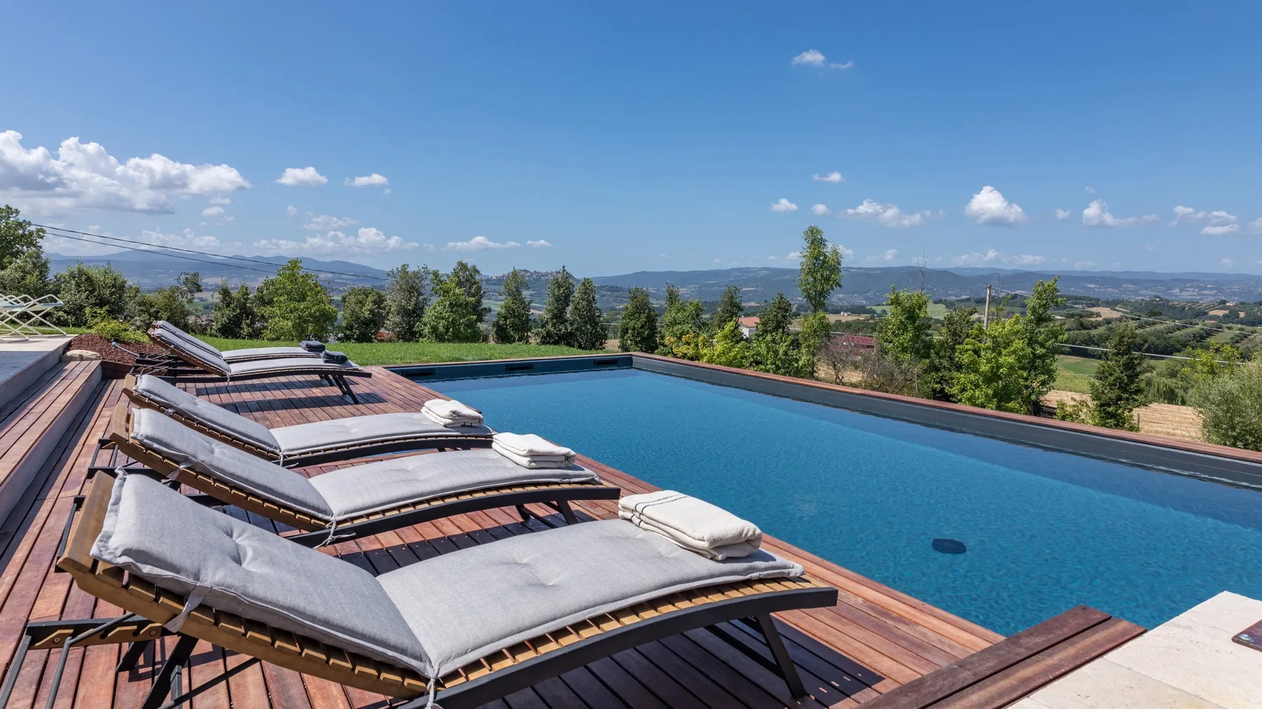 A rooftop pool sits beside wooden decking and cushioned loungers, overlooking a green landscape.