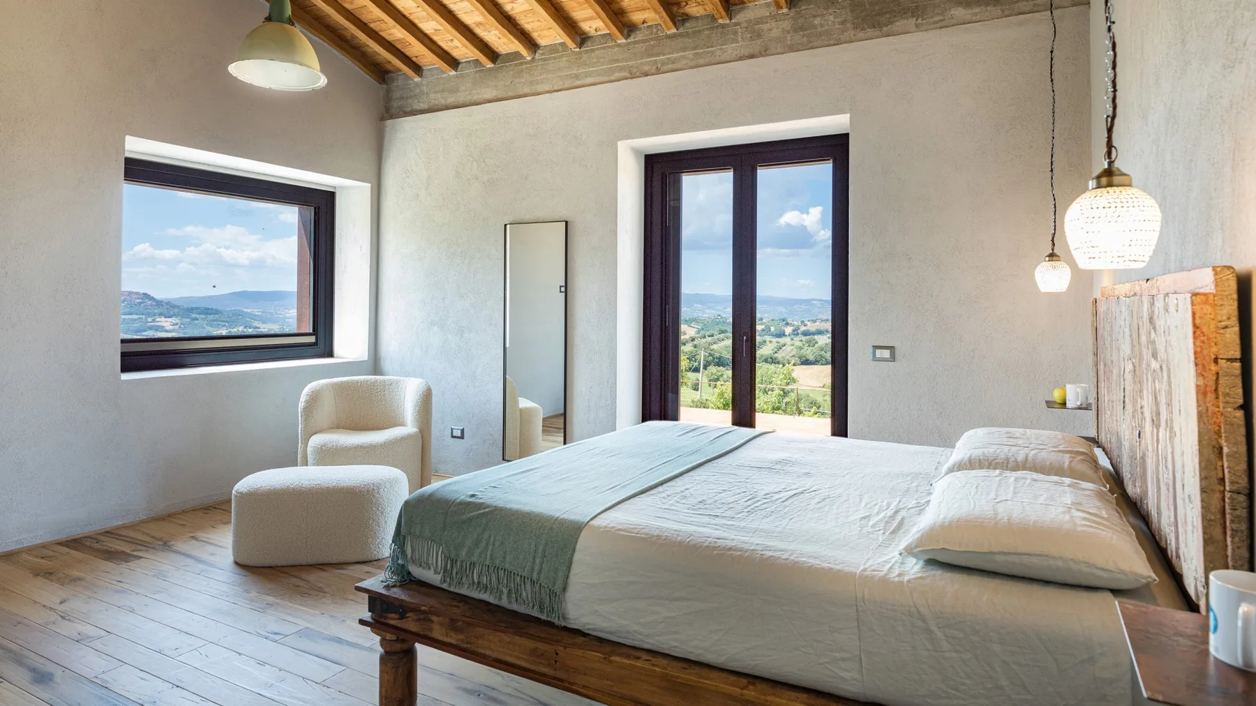 A bright bedroom has a wooden bed, pale bedding, a chair, and windows overlooking a green landscape.