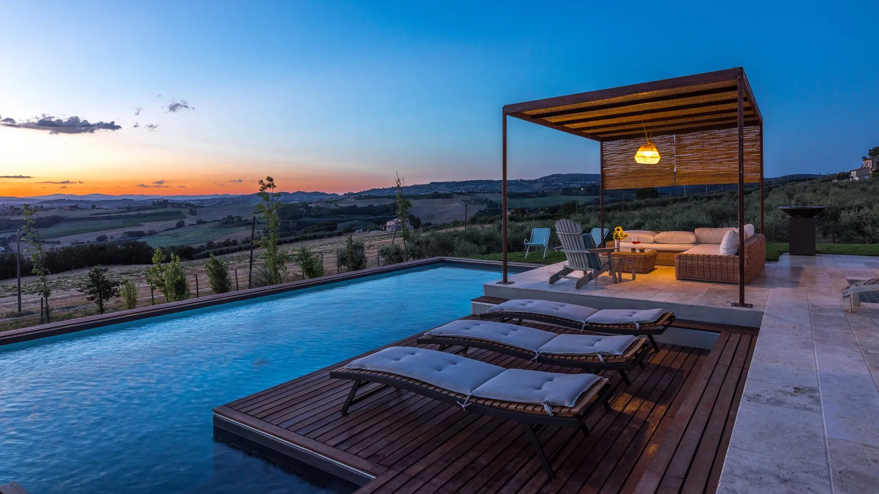 An infinity pool beside a wooden deck with lounge chairs and a covered seating area at sunset.
