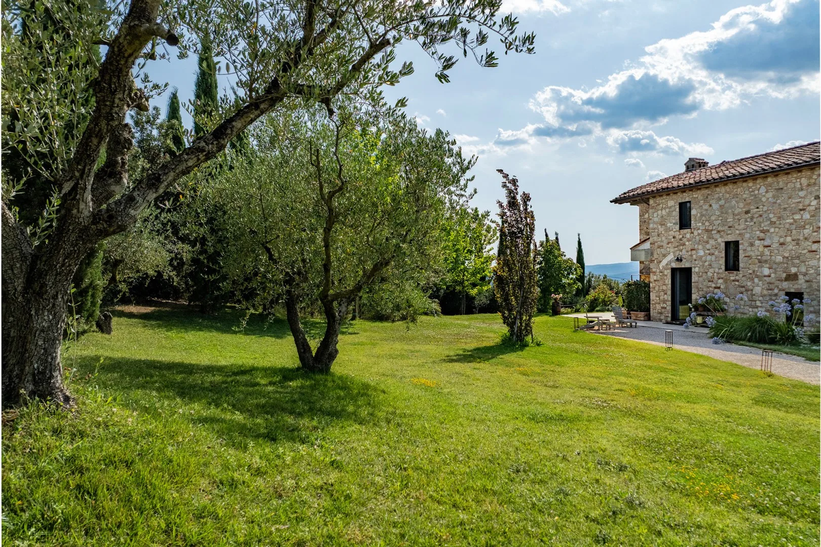 A stone house stands beside a broad green lawn, mature trees, and a paved walkway.