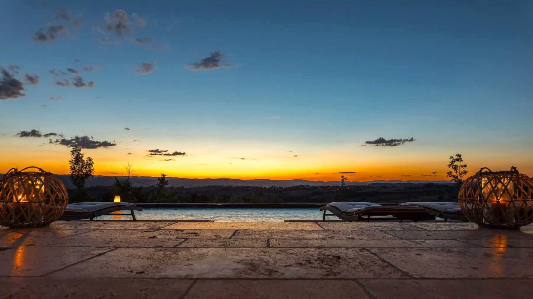 An outdoor pool and stone terrace overlook a distant landscape at sunset, with lounge chairs and decorative lanterns.