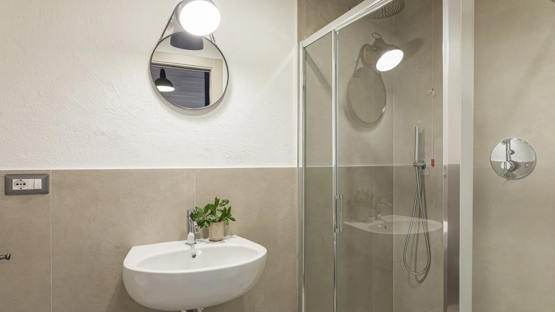 A bathroom with a wall-mounted sink, round mirror, and glass-enclosed shower.