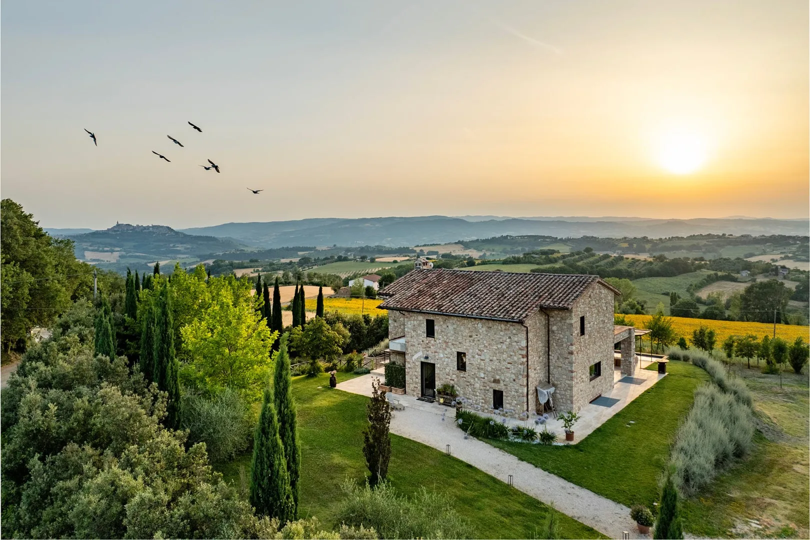 A stone farmhouse with a tiled roof stands among green fields and trees beneath a sunset sky.