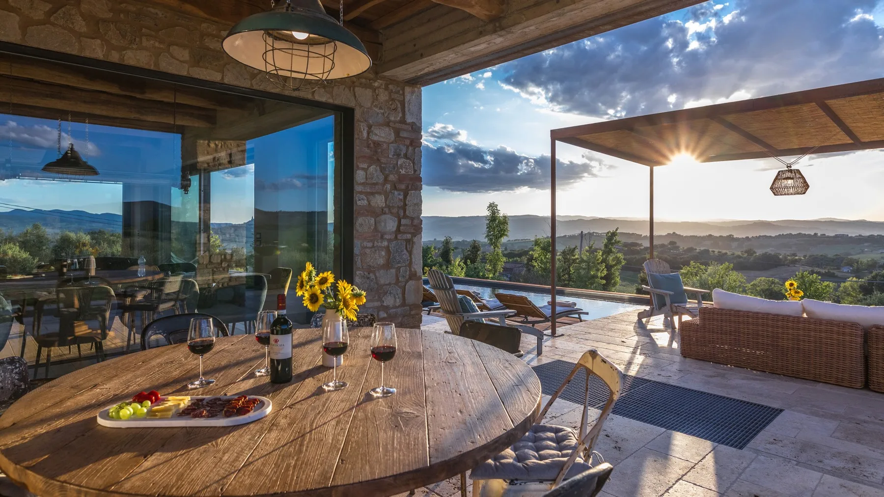 A stone-framed glass dining area opens onto a sunlit patio with a pool, outdoor seating, and distant hills.