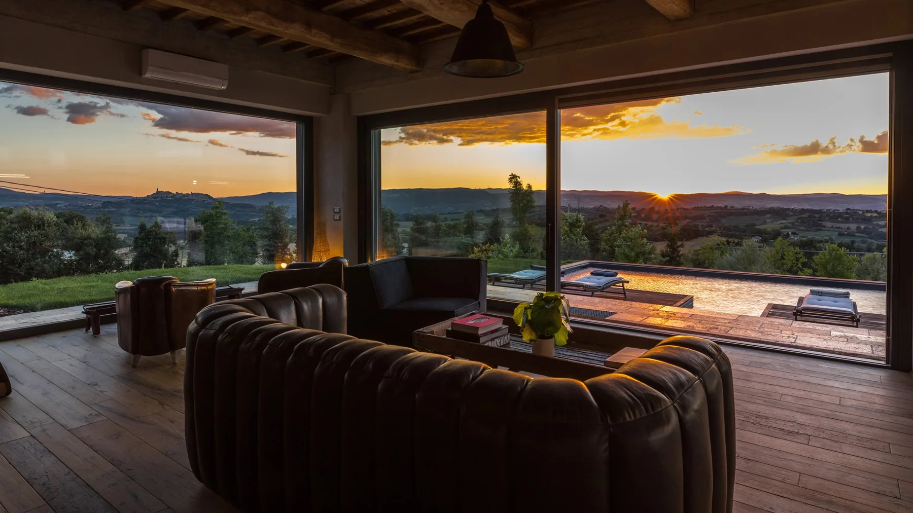 A living room with a dark leather sectional facing floor-to-ceiling windows and a sunset view.