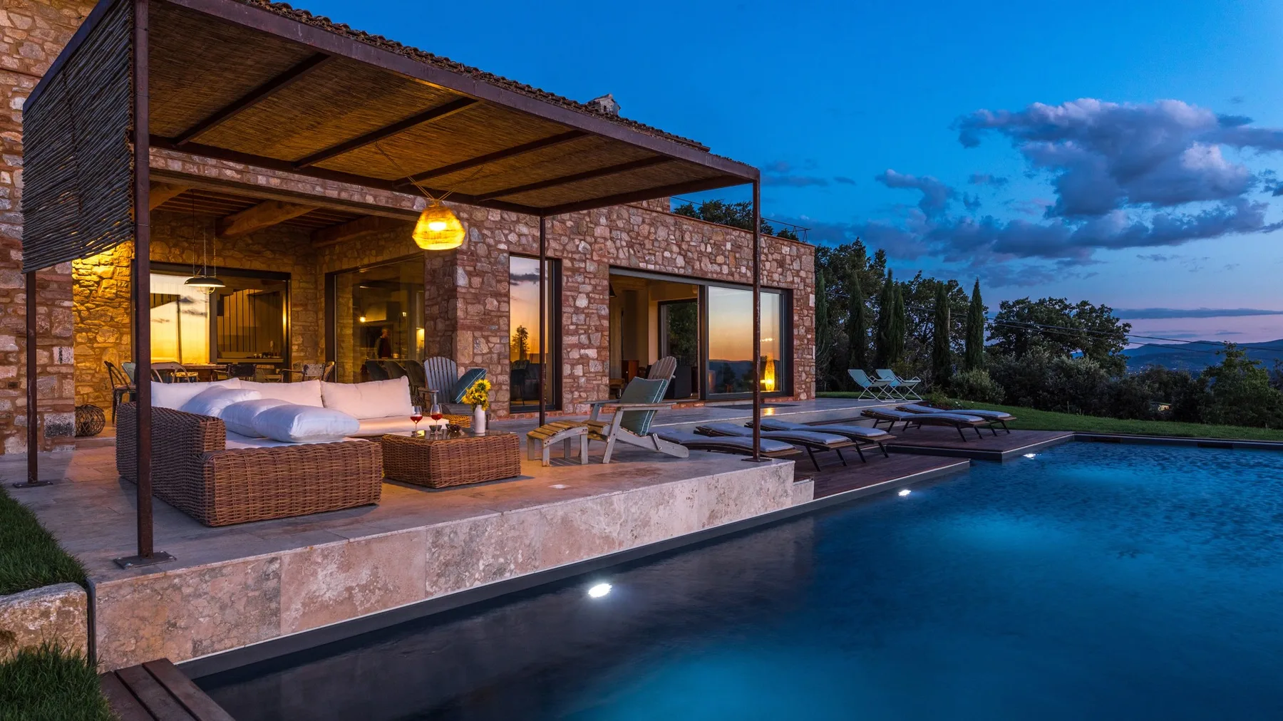 A modern stone villa opens onto a terrace with lounge seating beside an illuminated swimming pool at dusk.