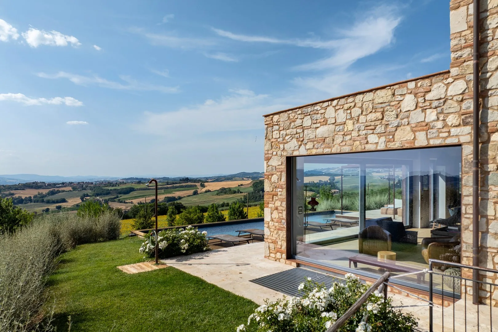 A stone house opens onto a patio and swimming pool, overlooking rolling countryside beneath a blue sky.