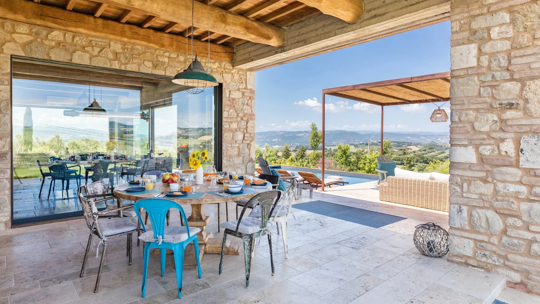 A stone terrace dining area has a set table, colorful metal chairs, and open views across the landscape.