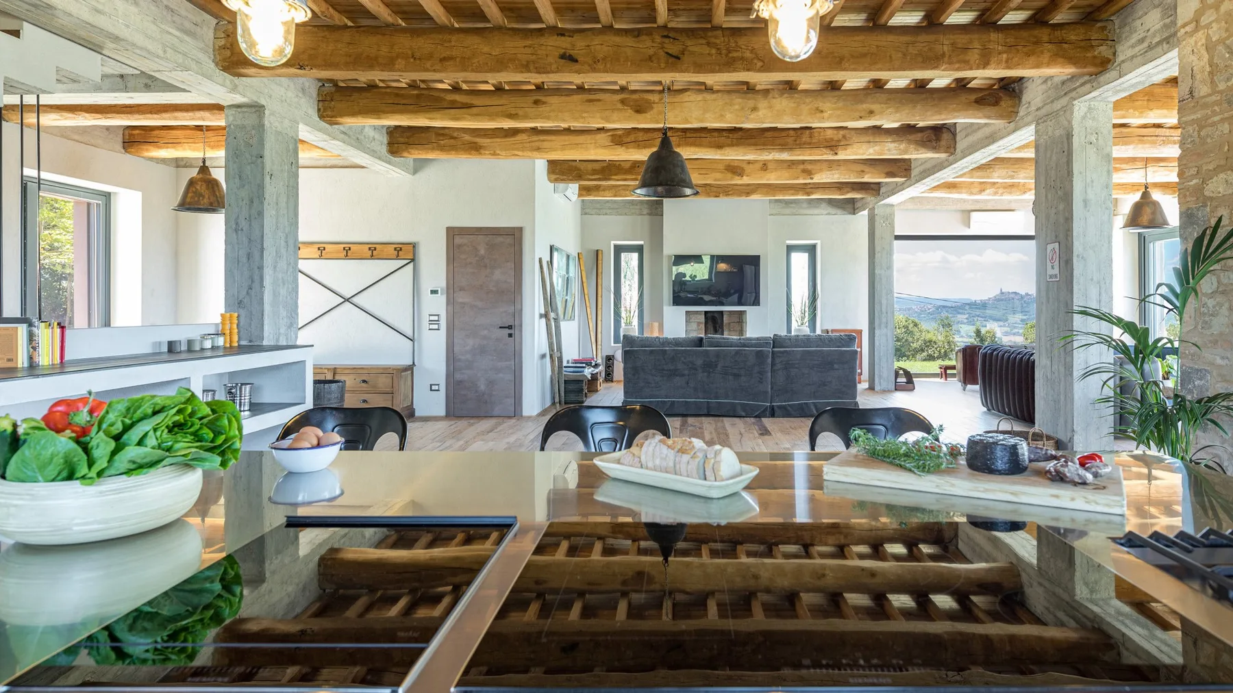 A bright open-plan living and dining area features a long glass table, exposed timber beams, and a sofa facing the television.