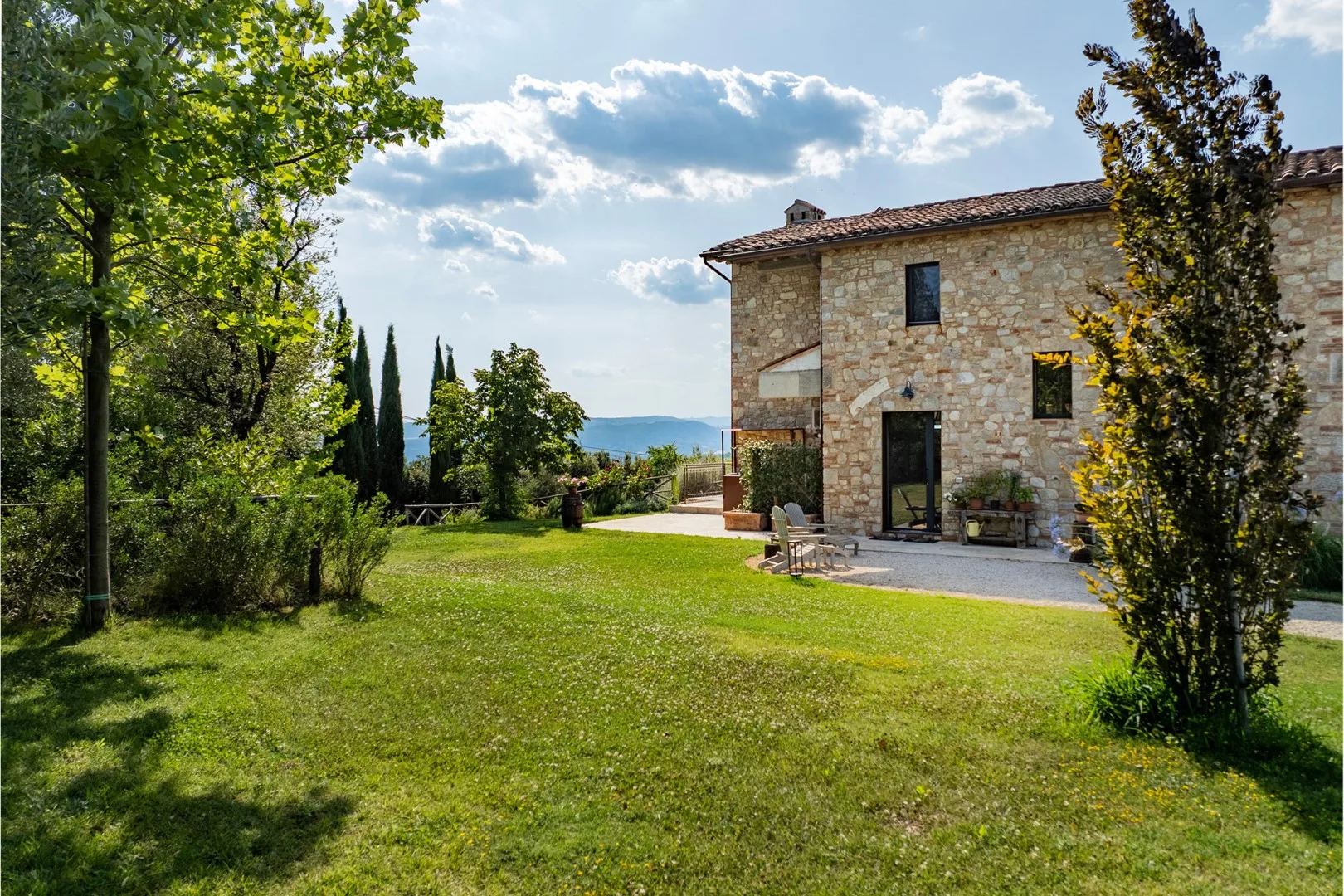 A stone house stands beside a broad green lawn, with leafy trees and a paved patio.
