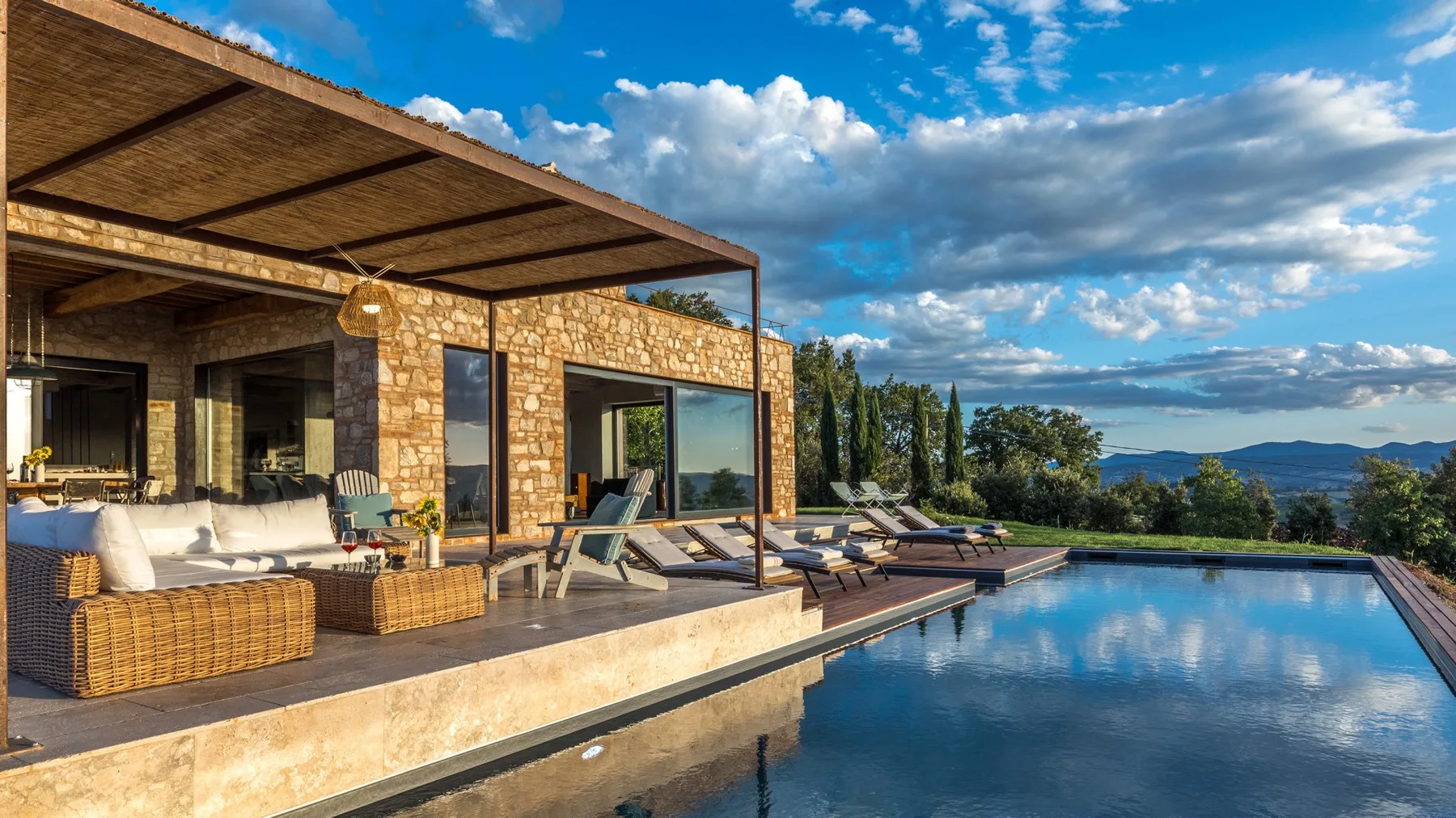 A stone house opens onto a poolside terrace with wicker seating, loungers, and a wide mountain view.
