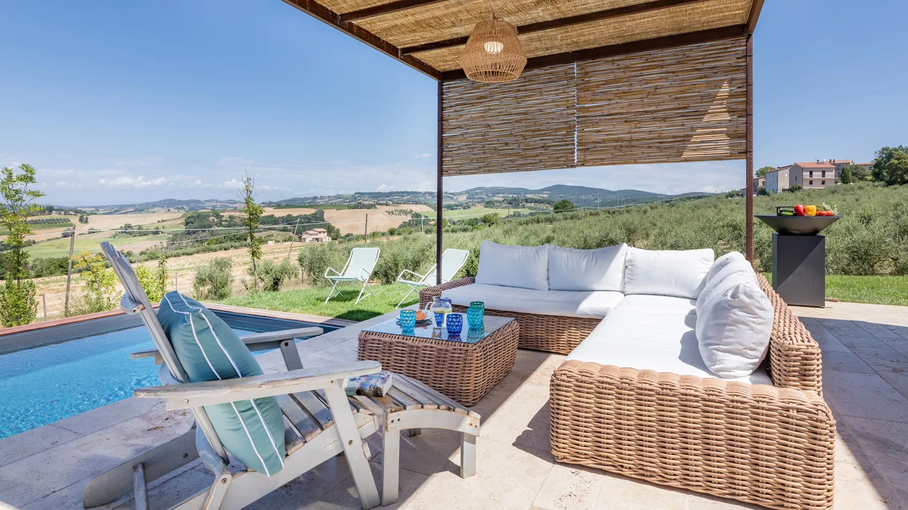A wicker sectional and lounge chairs sit beside a swimming pool on a sunny terrace overlooking fields.