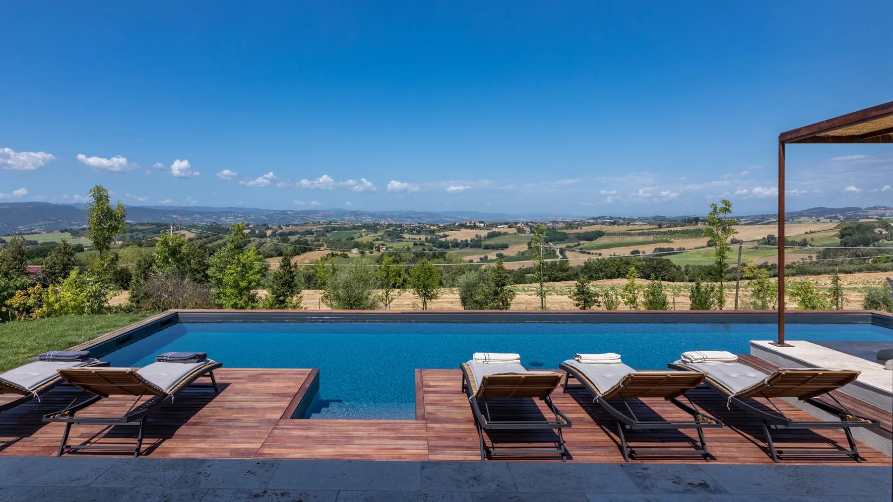 A swimming pool and wooden deck overlook a broad rural landscape beneath a clear blue sky.