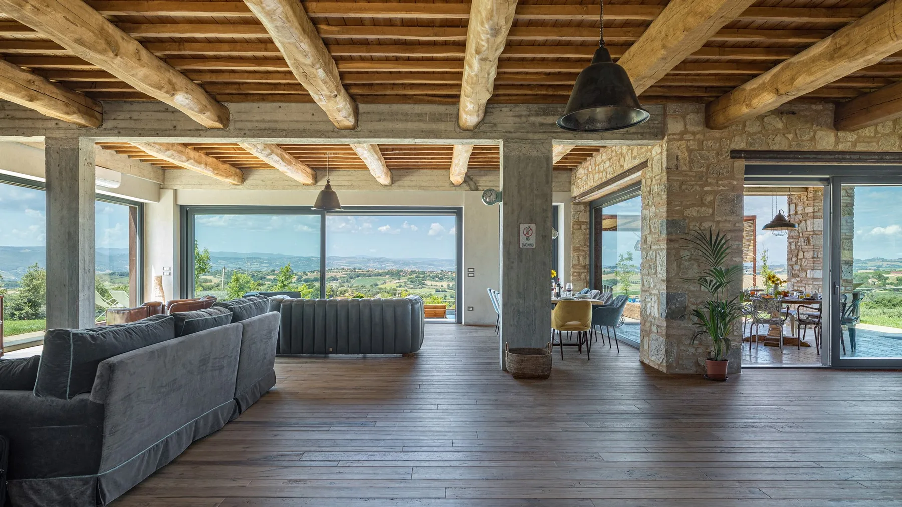 A spacious living room has a gray sectional, exposed timber beams, and wide windows overlooking a green landscape.