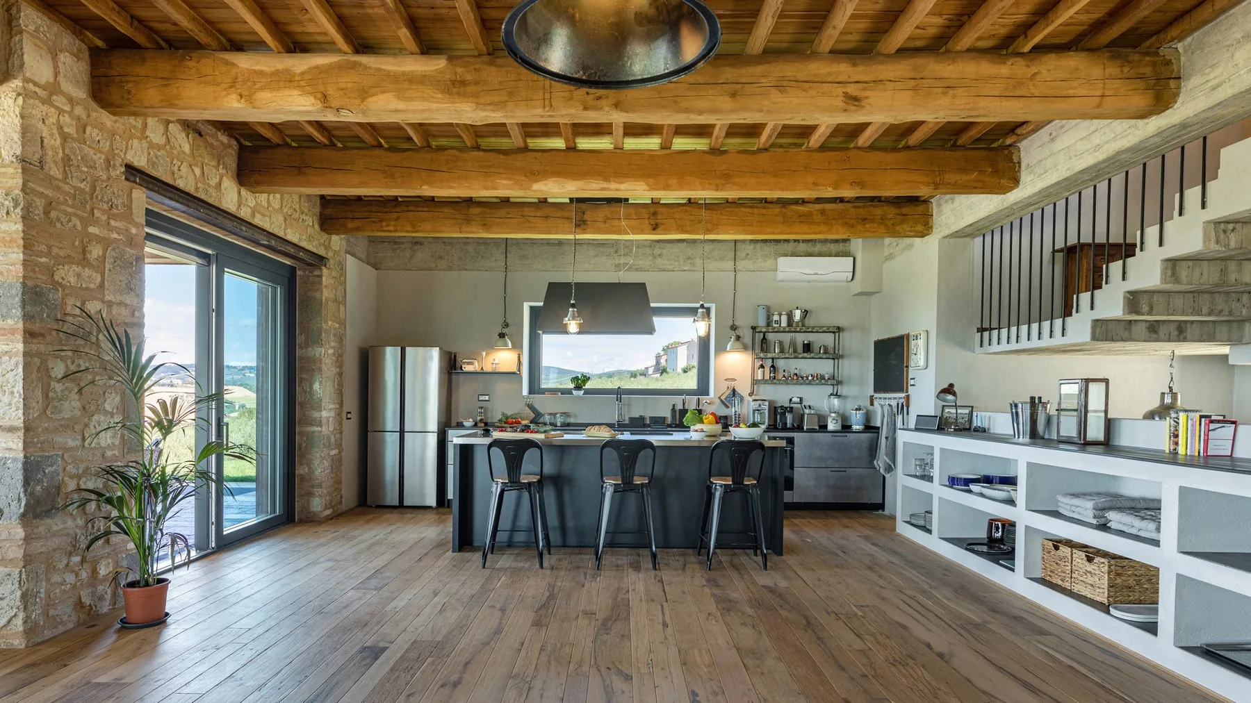 An open-plan kitchen and dining area features a long island, exposed wooden beams, and a stone-framed glass door.