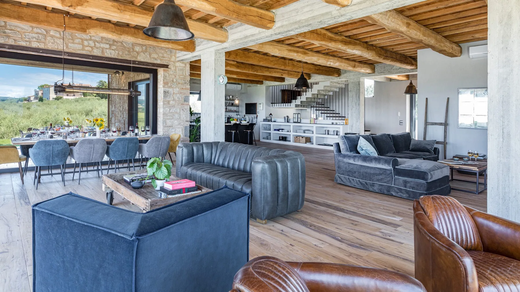 A spacious living and dining area features exposed timber beams, blue sofas, and a long table beside large windows.
