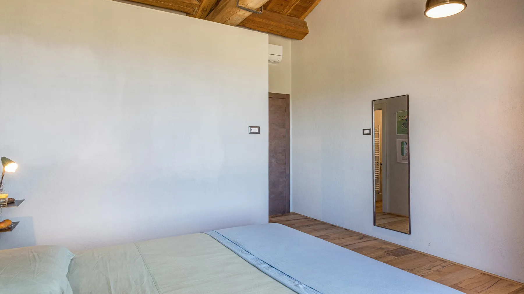 A bedroom with a low bed, pale blue bedding, exposed wooden ceiling beams, and a tall wall mirror.