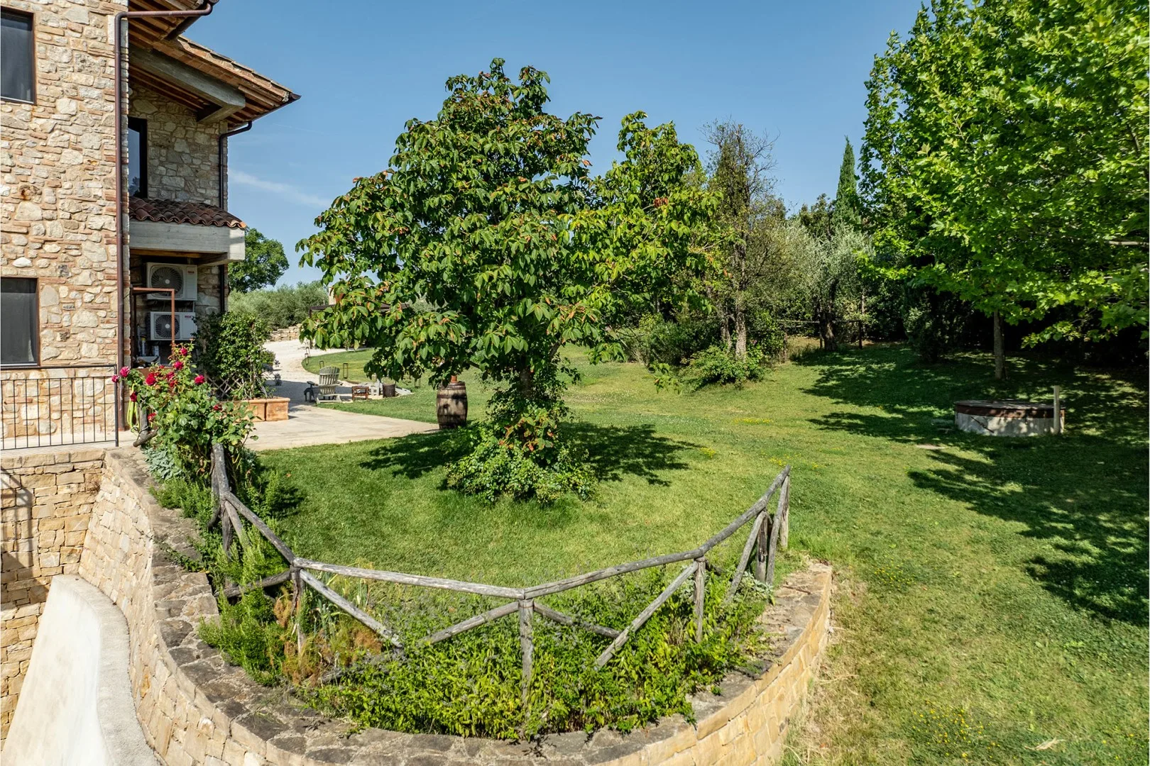 A stone house borders a sunny garden with a low wooden fence, leafy trees, and a grassy lawn.