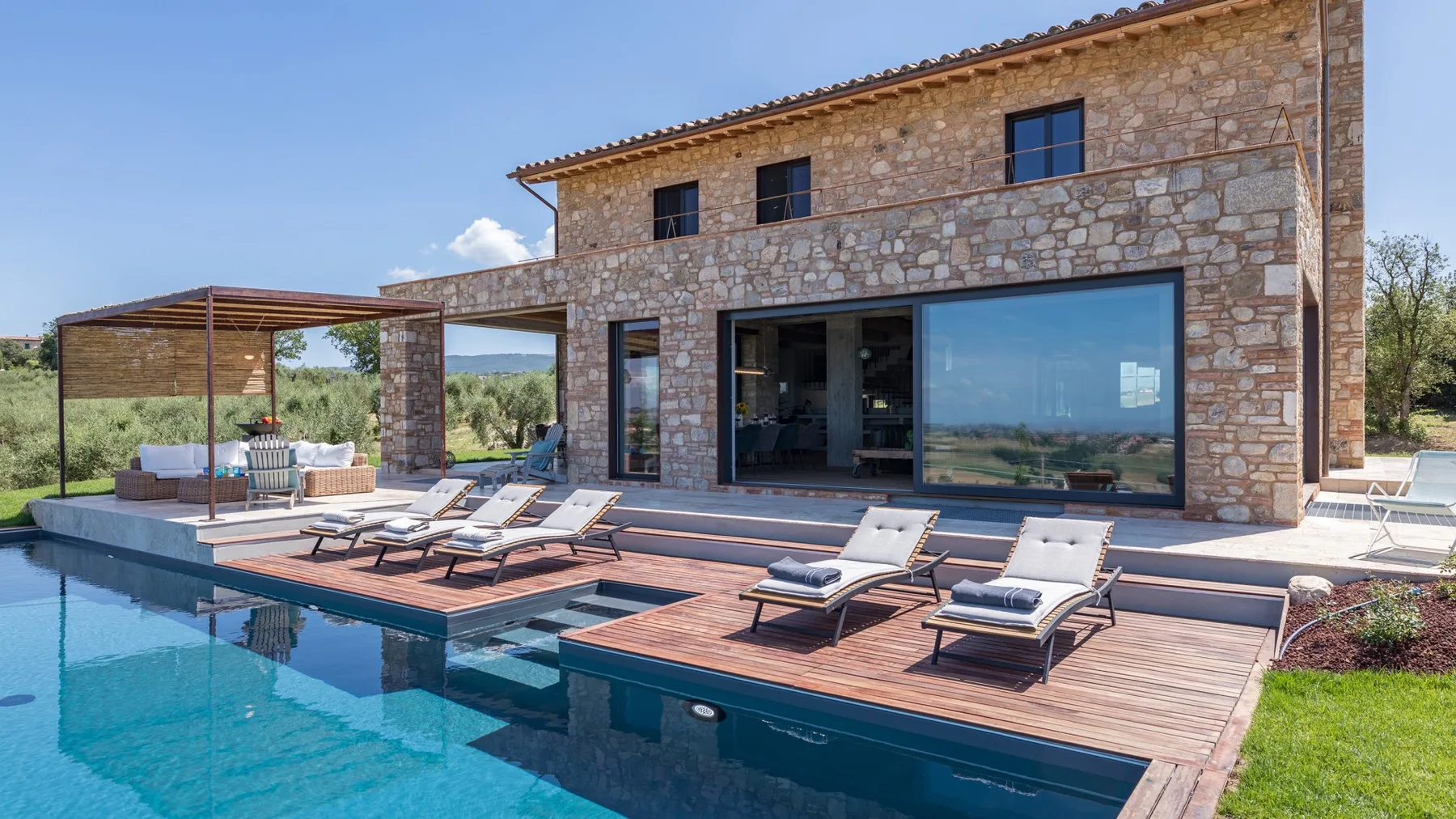 A stone house opens onto a swimming pool and wooden deck with several lounge chairs.
