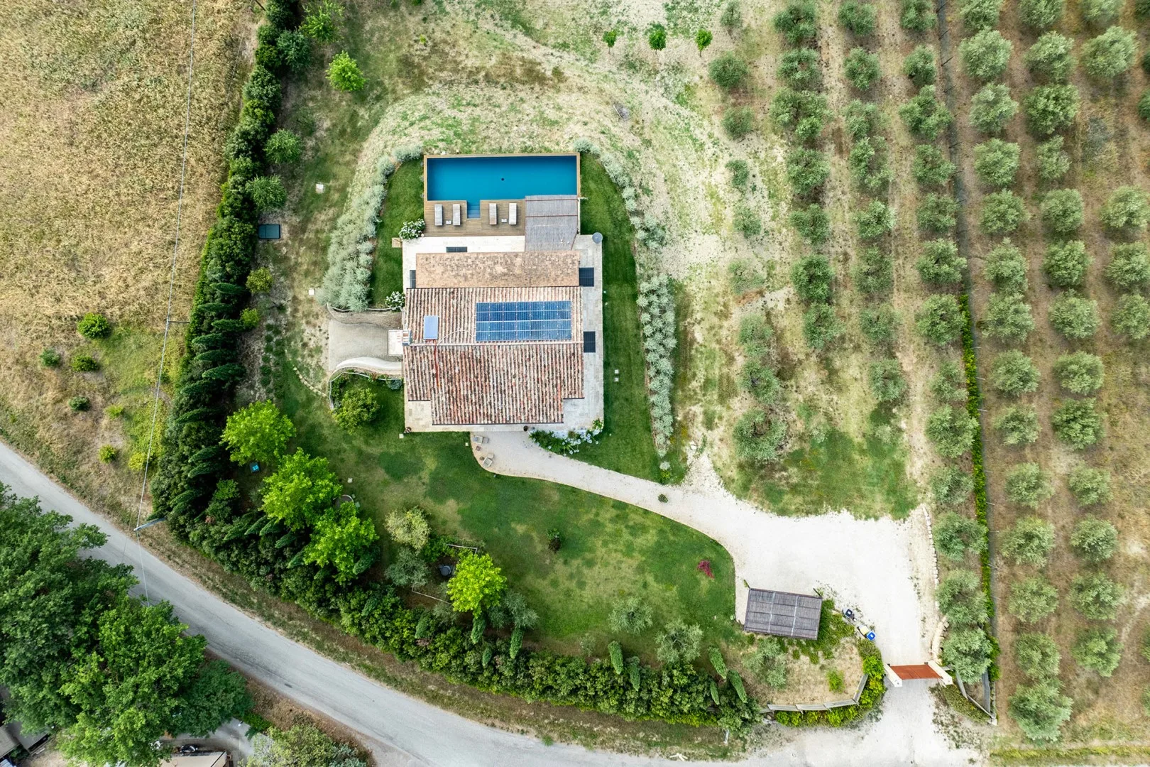 Aerial view of a tiled-roof house with a rectangular swimming pool, surrounded by cultivated fields and trees.